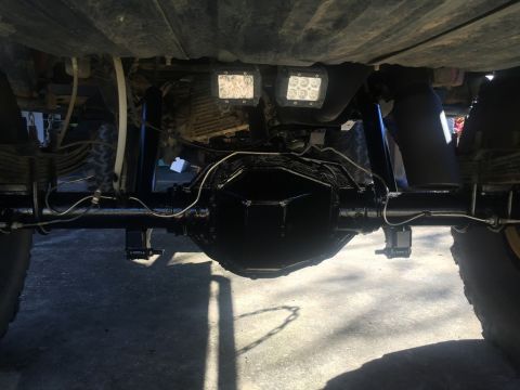 1989 Ford Bronco - The underside of a vehicle shows a black rear differential, leaf springs, and suspension components. Two square LED auxiliary lights are mo…
