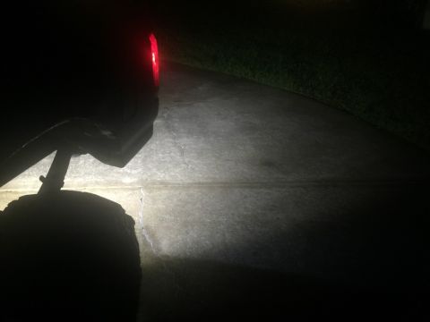 1989 Ford Bronco - The rear of a dark-colored vehicle is visible at night, with its red taillight illuminated. A bright light beam illuminates the concrete dr…