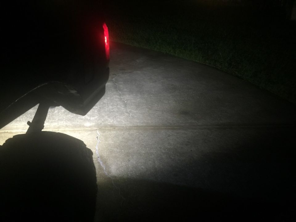 Rear of a dark vehicle at night with taillight on, illuminating a driveway and tire.