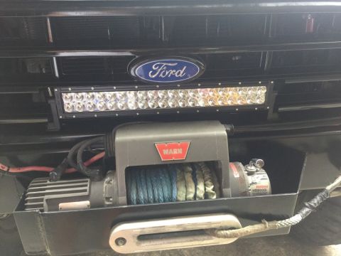 1989 Ford Bronco - A close-up front view of a black Ford Bronco's grille area shows a Ford emblem, a light bar, and a Warn winch mounted on a custom bumper.