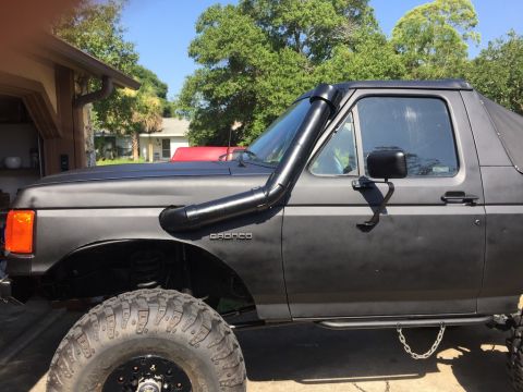 1989 Ford Bronco - The driver's side of a matte gray Ford Bronco is shown, featuring a snorkel intake mounted along the A-pillar and fender. The vehicle has l…