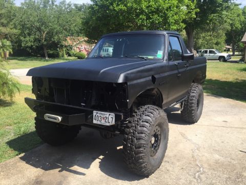 1989 Ford Bronco - This modified pickup truck has large off-road tires and a matte black finish. Another vehicle is visible in the background.