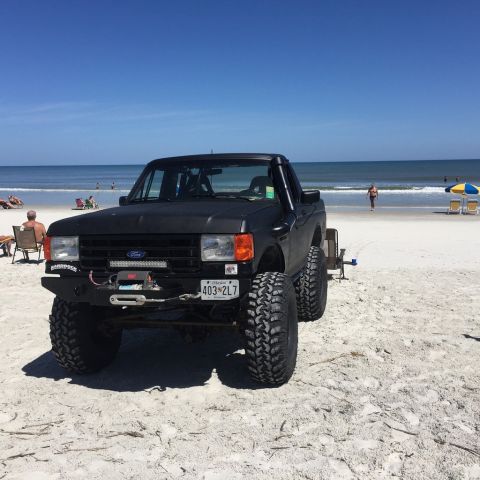 1989 Ford Bronco - A black Ford Bronco with large off-road tires is parked on a sandy beach with the ocean and a few people in the background.