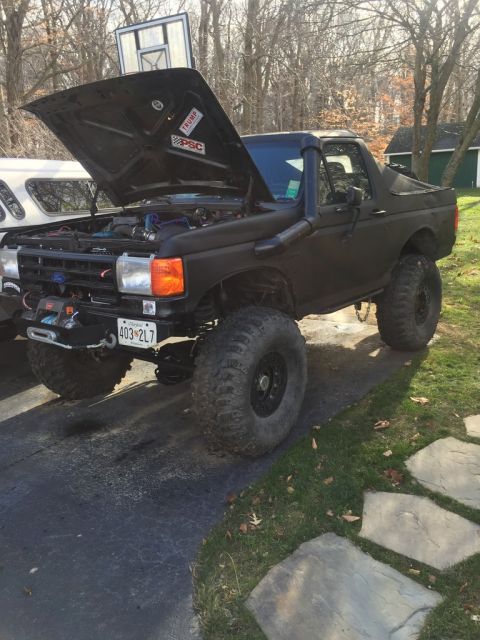 1989 Ford Bronco - A matte black Ford Bronco with the hood open is parked on a driveway. The vehicle has large off-road tires, a snorkel, and a winch mounted…