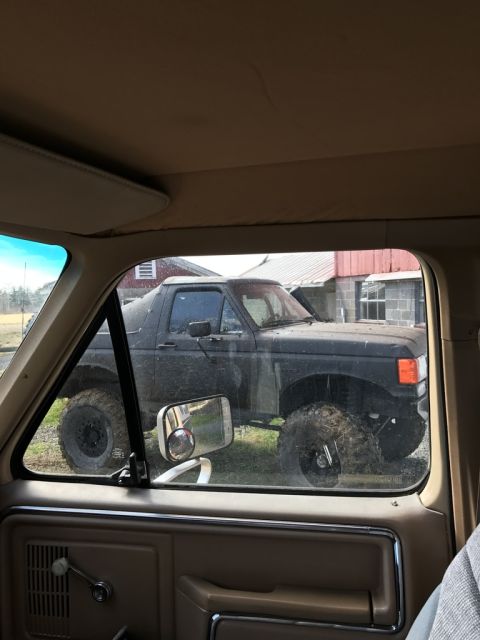 1989 Ford Bronco - View from inside a vehicle looking out the driver's side window at a lifted, dark-colored Ford Bronco with large, muddy tires. The Bronco i…