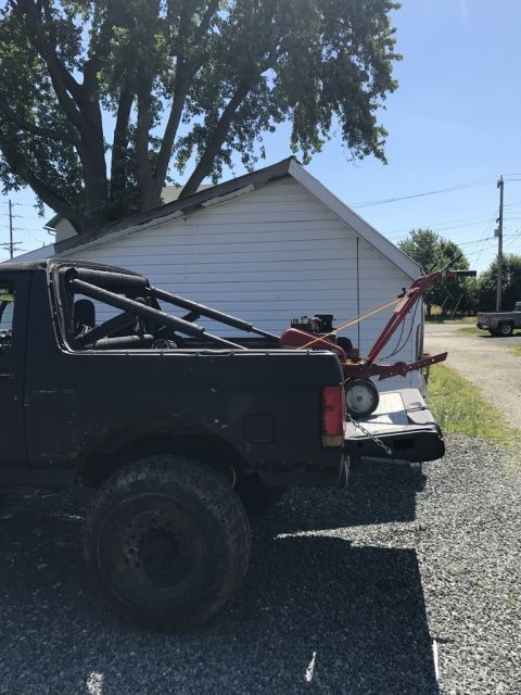 1989 Ford Bronco - A black Ford Bronco with a roll cage is parked on a gravel lot next to a white building. A red tiller is loaded in the bed of the truck.