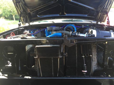 1989 Ford Bronco - The open hood of a black Ford Bronco reveals its engine bay and front grille assembly. Visible modifications include bright blue hoses and…