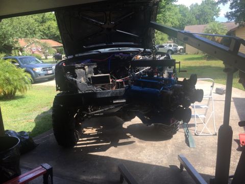 1989 Ford Bronco - A large engine is being hoisted into the engine bay of a black Ford Bronco with its hood open. A blue SUV is parked in the background.