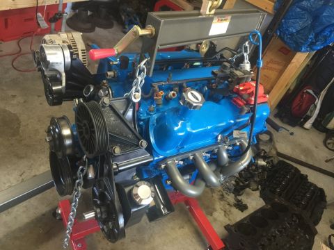 1989 Ford Bronco - A blue V8 engine is mounted on a red engine stand, with an alternator and various pulleys visible on the front. The engine is suspended by…
