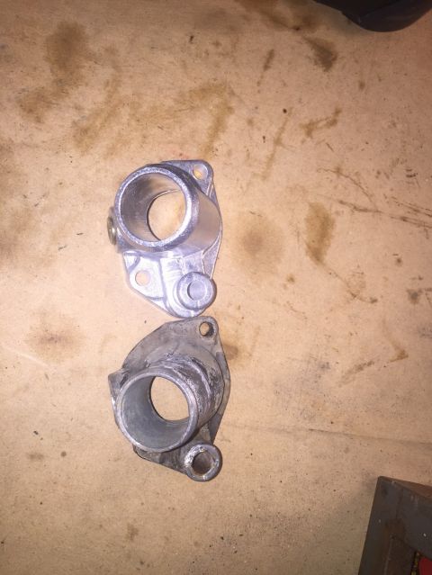 1989 Ford Bronco - Two metal automotive parts, likely thermostat housings, are shown on a brown, stained surface. The upper part is clean and silver, while th…