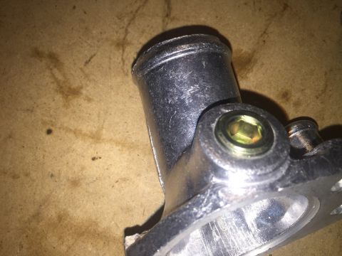 1989 Ford Bronco - A close-up view shows a metal automotive part, likely a thermostat housing, with a hex bolt secured in the center. The part is silver and h…