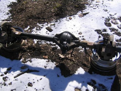 1978 Ford Bronco - A bare front axle assembly lying on patchy snow and dirt, photographed from above with its offset cast pumpkin, tubes, knuckles, disc brake…