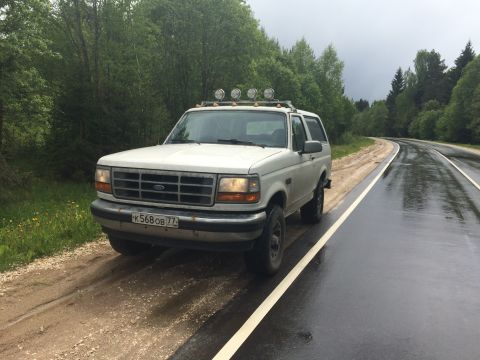 1993 Ford Bronco - White 1993 Ford Bronco sits on the side of a wet road surrounded by trees.