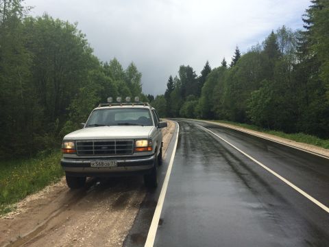 1993 Ford Bronco - A 1993 Ford Bronco is parked on the shoulder of a wet road surrounded by lush green trees.