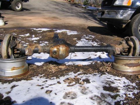 1978 Ford Bronco - A rusty bare front axle assembly resting on two old steel wheels in a snow-patched driveway, shown head-on with its offset differential hou…