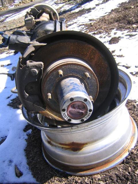 1978 Ford Bronco - Close-up of a front axle end resting on a bare chrome/aluminum wheel outdoors in patchy snow, showing the rusty vented brake rotor, black c…