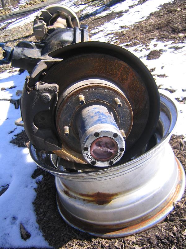 Front axle end with rusty brake rotor, caliper and manual locking hub sitting on a wheel in the snow