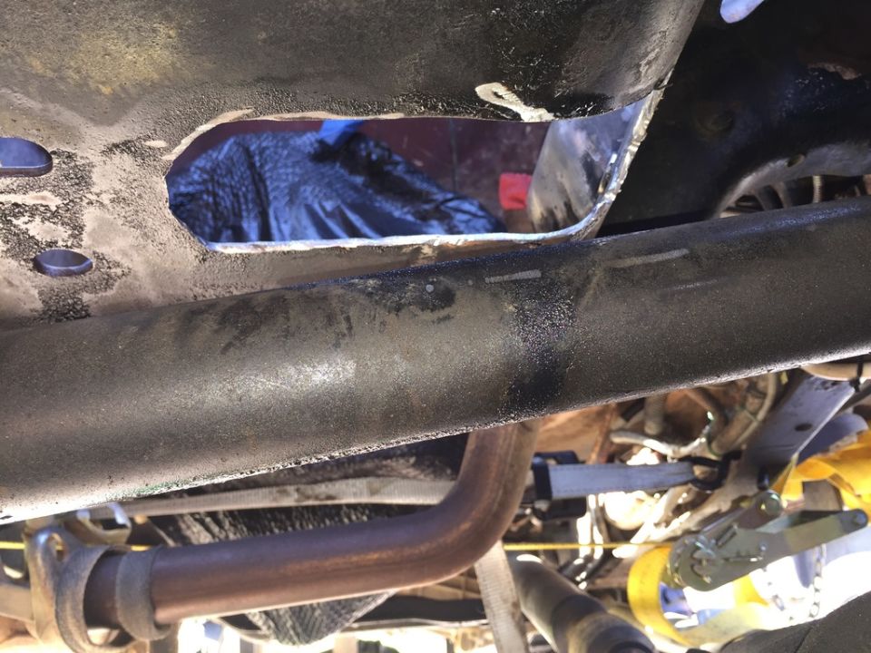 Close-up of a vehicle's underbody showing a large hole cut into the frame rail. Exhaust pipe visible below