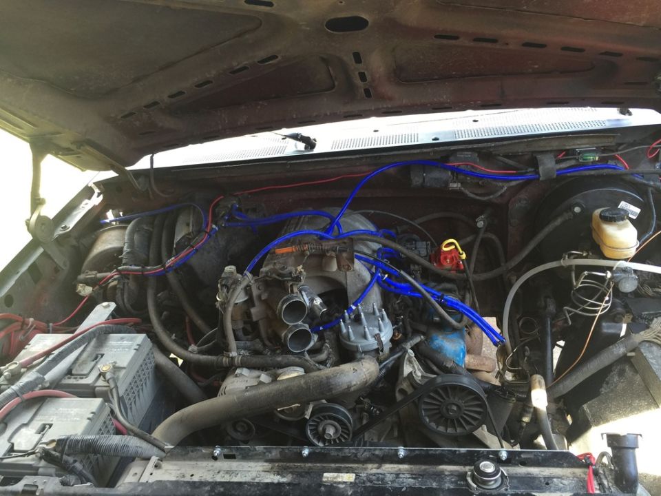 Engine bay showing a V8 engine with blue spark plug wires, distributor, and intake manifold. Batteries are visible on the left