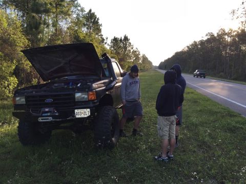 1989 Ford Bronco - A group of people stand around a black Ford truck with its hood open, parked on the side of a road.