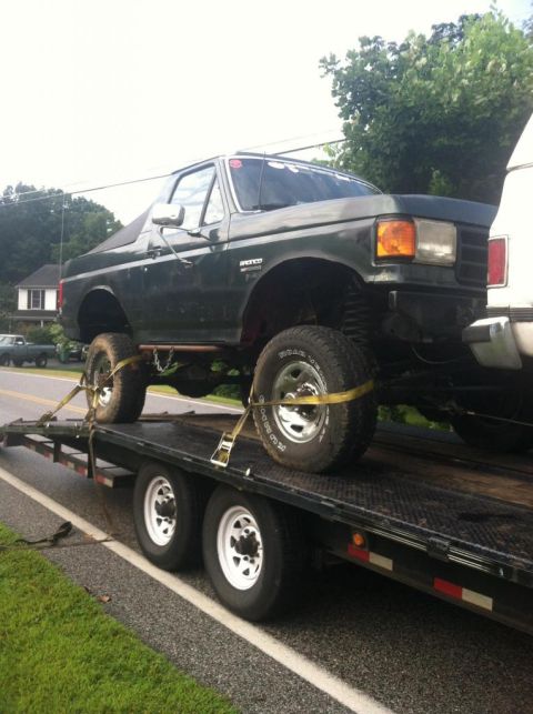 1989 Ford Bronco - A lifted Ford Bronco is secured with straps on a flatbed trailer, ready for transport.