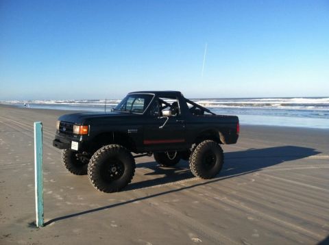 1989 Ford Bronco - A lifted, dark-colored Ford Bronco with large off-road tires is parked on a sandy beach with the ocean in the background. The vehicle has a…