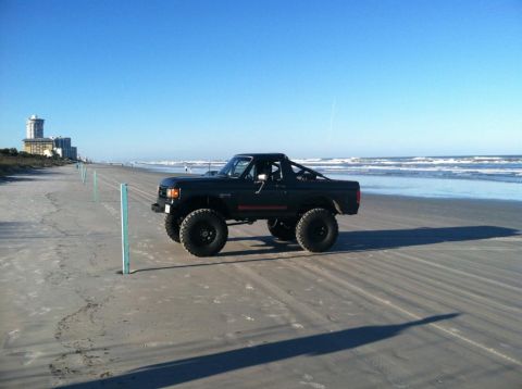 1989 Ford Bronco - A lifted, dark green Ford Bronco is parked on a sandy beach with the ocean and buildings in the background.