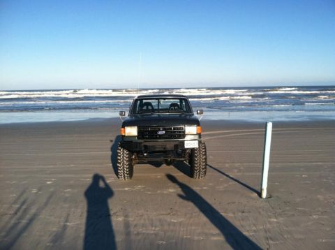 1989 Ford Bronco - A lifted, dark colored Ford truck is parked on a sandy beach facing the ocean. The truck has large, aggressive tires and a custom front bum…
