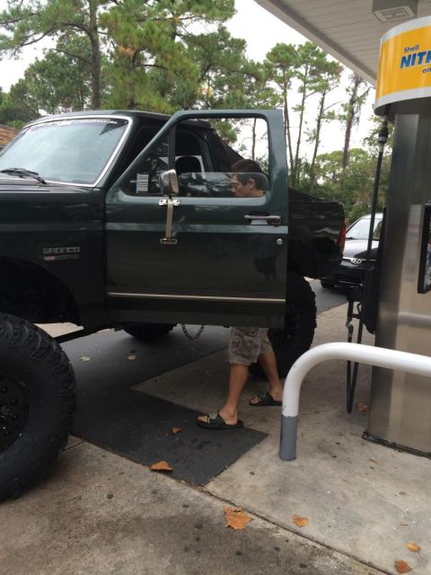 1989 Ford Bronco - The driver's side of a dark green Ford Bronco is shown with the door open at a gas station. A person wearing shorts and sandals is standing…