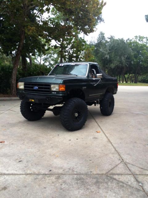 1989 Ford Bronco - A dark green, lifted 1989 Ford Bronco with large off-road tires is parked on a concrete surface with trees in the background.