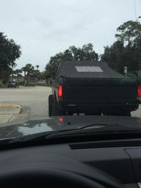 1989 Ford Bronco - The rear view of a black Ford Bronco with a soft top is seen from inside another vehicle on a rainy day. The Bronco has large tires and red…