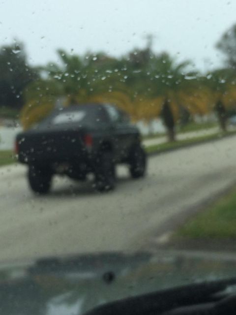 1989 Ford Bronco - The rear of a black Ford Bronco is visible through a rain-streaked windshield, driving down a residential street with palm trees in the bac…