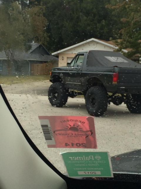 1989 Ford Bronco - A lifted black Ford Bronco with a soft top is parked on a wet street. Two stickers are visible on the windshield, one red and one green.