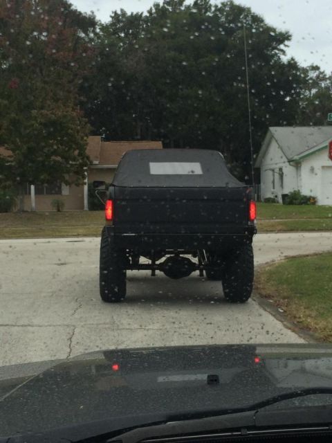 1989 Ford Bronco - The rear view of a black, lifted Ford Bronco with a soft top is shown on a wet road. The vehicle has large off-road tires and visible suspe…