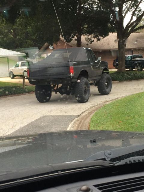 1989 Ford Bronco - A lifted, dark-colored Ford Bronco with a black soft top is parked on a suburban street. The vehicle features oversized tires and appears t…