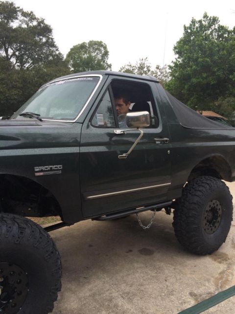 1989 Ford Bronco - Side view of a dark green Ford Bronco with large off-road tires and a black soft top. A man is visible through the driver's side window.