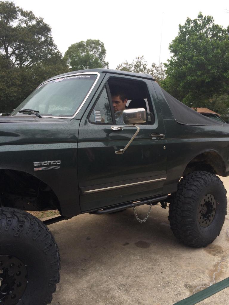Dark green Ford Bronco with large tires and a black soft top, driver visible.