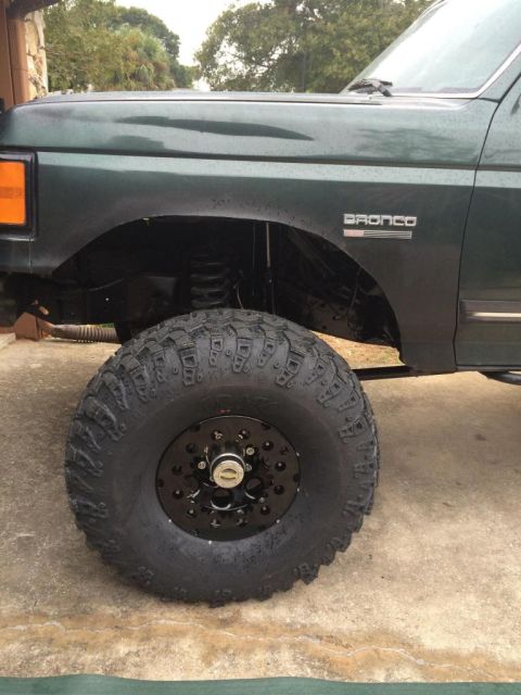 1989 Ford Bronco - A close-up, low-angle shot of the front driver's side fender and oversized tire of a dark green Ford Bronco. The vehicle has a lifted suspe…