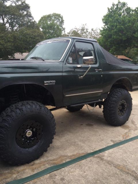 1989 Ford Bronco - A dark green Ford Bronco is parked on a concrete surface, showing its large off-road tires and lifted suspension. The vehicle has a soft to…