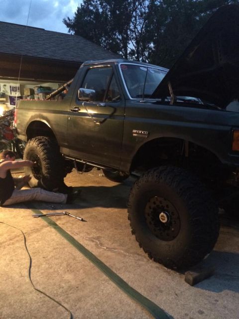 1989 Ford Bronco - A dark green Ford Bronco is parked on a concrete driveway with its hood open. A person is lying on the ground near the front wheel, appeari…