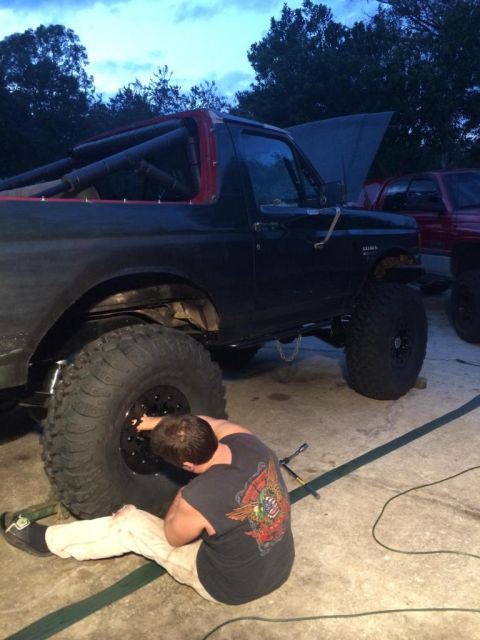 1989 Ford Bronco - A person works on the front tire of a black Ford Bronco with a roll cage visible in the bed. The vehicle is parked on a concrete surface wi…