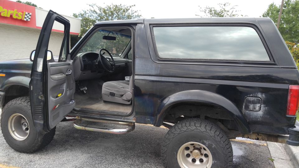 Black 1996 Ford Bronco with driver's door open, parked outside a parts store.
