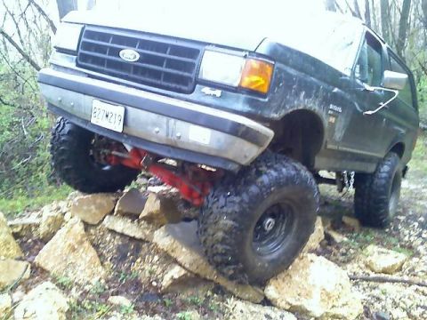 1989 Ford Bronco - A dark-colored Ford Bronco is shown from a low angle, driving over large rocks on a dirt path. The front of the vehicle is angled towards t…