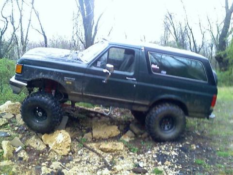 1989 Ford Bronco - A dark green 1989 Ford Bronco is shown from the side, lifted with large off-road tires, parked on a rocky incline with trees in the backgro…
