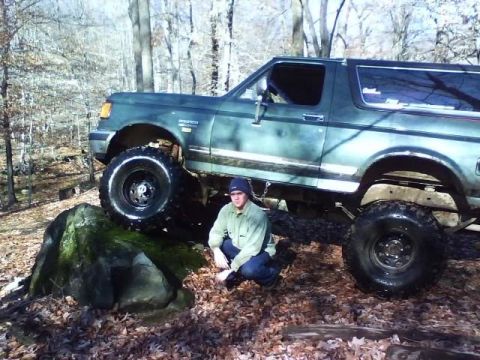 1989 Ford Bronco - A man kneels in front of a lifted, dark green Ford Bronco that is partially on a large moss-covered rock in a wooded area with fallen leave…