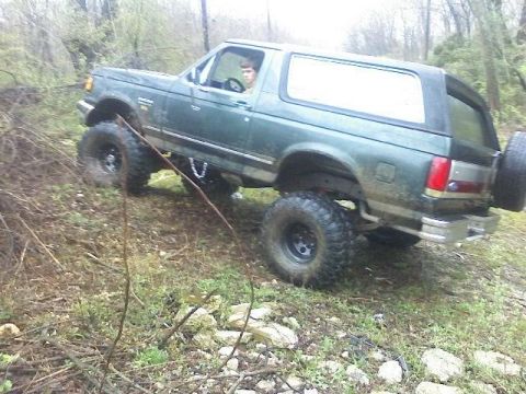 1989 Ford Bronco - A lifted, dark green 1989 Ford Bronco is shown off-road, navigating a muddy incline. The vehicle features large, aggressive tires and appea…