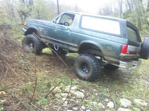1989 Ford Bronco - A dark green Ford Bronco is shown from the side, angled uphill on a dirt path with trees and brush in the background. The vehicle has large…