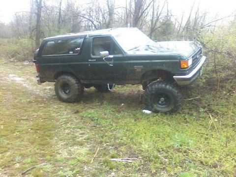 1989 Ford Bronco - A dark green, lifted Ford Bronco is parked on grassy terrain with trees in the background. The vehicle has large off-road tires and appears…