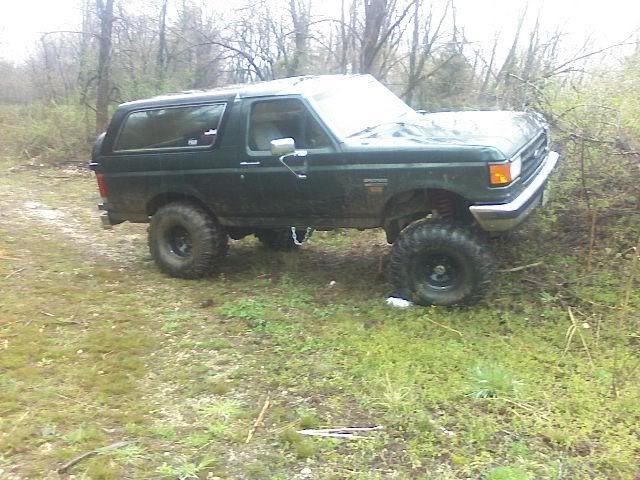1980s Ford Bronco with lift kit and off-road tires parked in a grassy, wooded area.