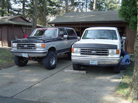1989 Ford Bronco - Two Ford trucks, a lifted Bronco and a pickup, are parked in a driveway next to a garage.